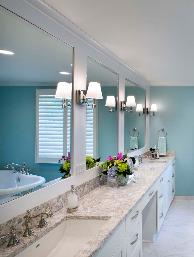Double sink bathroom vanity with marble countertop, bright lighting, and decorative floral arrangeme.