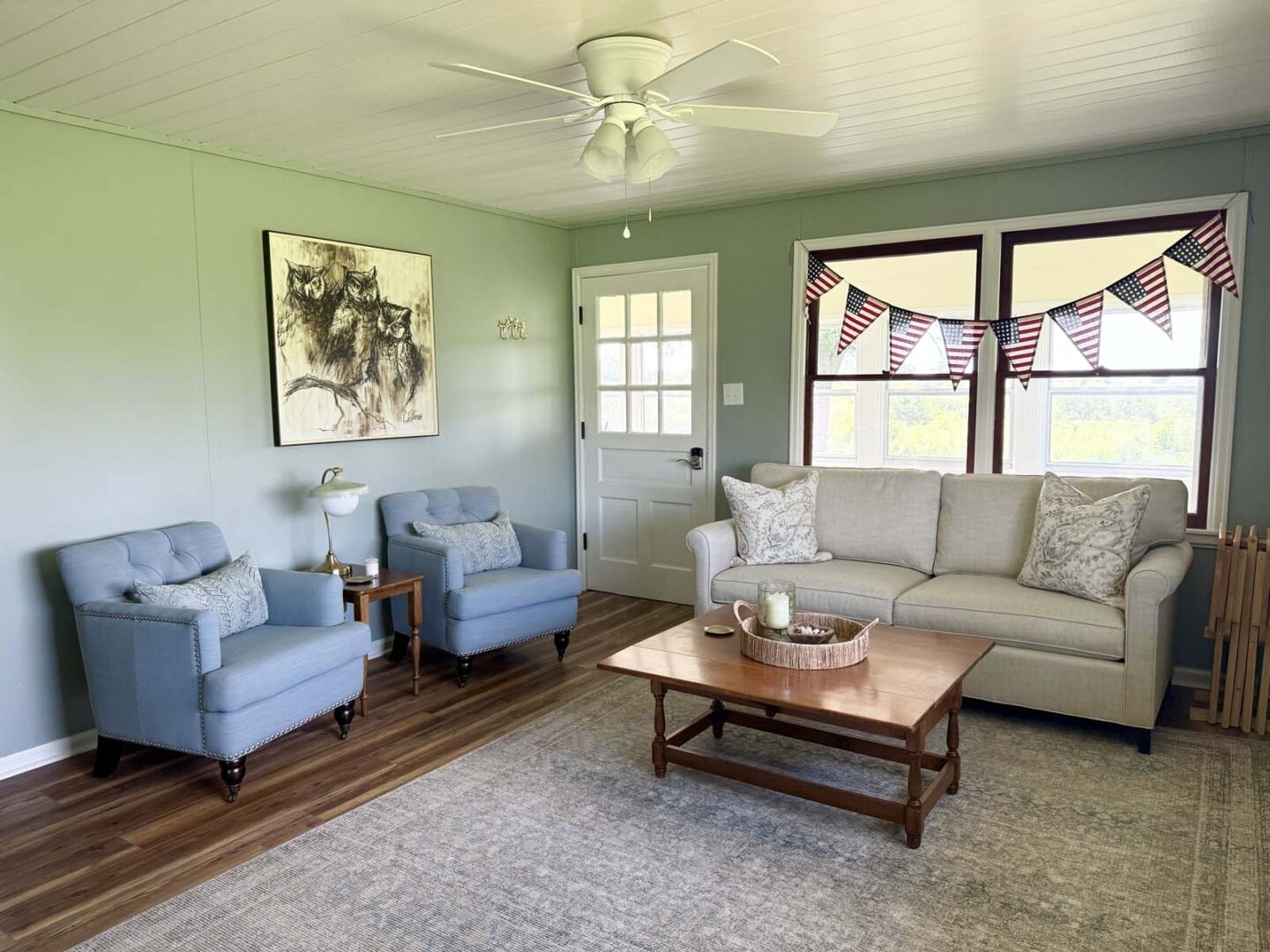 Bright living room featuring a beige sofa, blue armchairs, and patriotic bunting by the window.